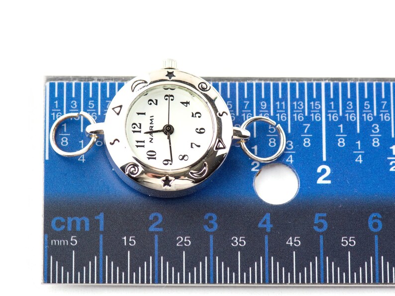 May include: A silver-tone round watch face with a white dial, black numerals, and the text NARMI. The bezel features engraved stars, moons, and spirals. Two silver rings are attached to the sides. A ruler shows the watch is 1 inch in diameter.