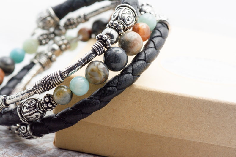 May include: A black leather bracelet with silver accents and multi-colored gemstone beads. The bracelet is wrapped around a brown box.