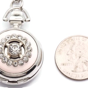 May include: A silver locket with a pink enamel background and a crystal center surrounded by smaller crystals. The locket is about the size of a quarter.