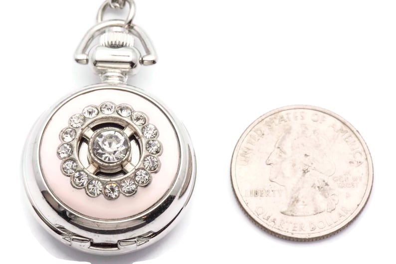 May include: A silver locket with a pink enamel background and a crystal center surrounded by smaller crystals. The locket is about the size of a quarter.