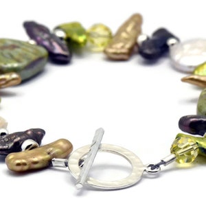 K&ouml;nnte beinhalten: Ein Armband aus gr&uuml;nen, braunen und violetten Perlen mit einem silbernen Schiebeverschluss. Das Armband besteht aus verschiedenen unregelm&auml;&szlig;ig geformten Perlen.