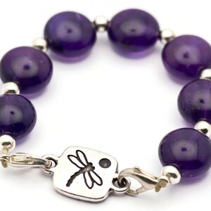 May include: A purple amethyst gemstone bracelet with a silver dragonfly charm. The bracelet is made with round beads and a silver clasp.