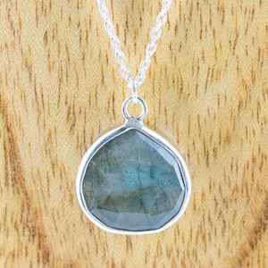 Pu&ograve; includere: Una collana d'argento con un ciondolo a forma di goccia. Il ciondolo presenta una pietra preziosa blu-verde iridescente e sfaccettata incastonata in una lunetta d'argento. La catena &egrave; una delicata maglia d'argento. Lo sfondo &egrave; una venatura del legno marrone chiaro.