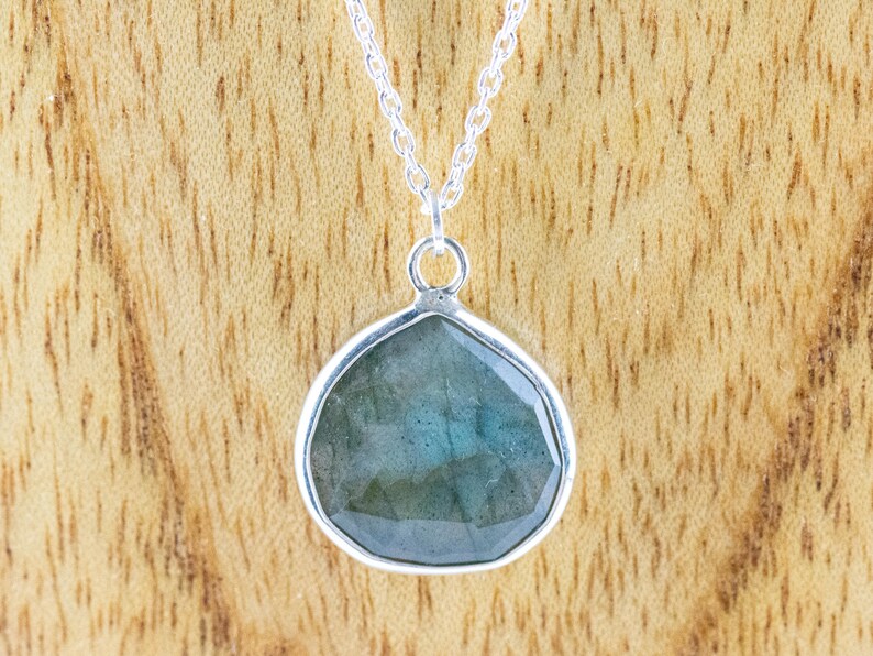 Pu&ograve; includere: Una collana d'argento con un ciondolo a forma di goccia. Il ciondolo presenta una pietra preziosa blu-verde iridescente e sfaccettata incastonata in una lunetta d'argento. La catena &egrave; una delicata maglia d'argento. Lo sfondo &egrave; una venatura del legno marrone chiaro.