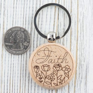 May include: Wooden keyring with a silver key ring. The keyring is round and has the word "Faith" engraved on it. The word is surrounded by a floral design.