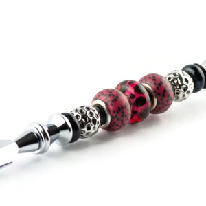 May include: A decorative bottle stopper with a silver metal handle and a series of beads. The beads include red and black patterned spheres, silver textured beads, and black and silver accents. The stopper is designed for a bottle.
