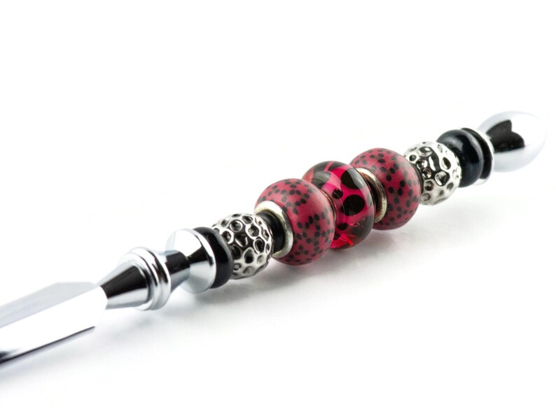 May include: A decorative bottle stopper with a silver metal handle and a series of beads. The beads include red and black patterned spheres, silver textured beads, and black and silver accents. The stopper is designed for a bottle.