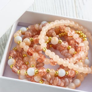 May include: A set of six bracelets made with pink, peach, and white beads. The bracelets are stacked inside a white gift box.