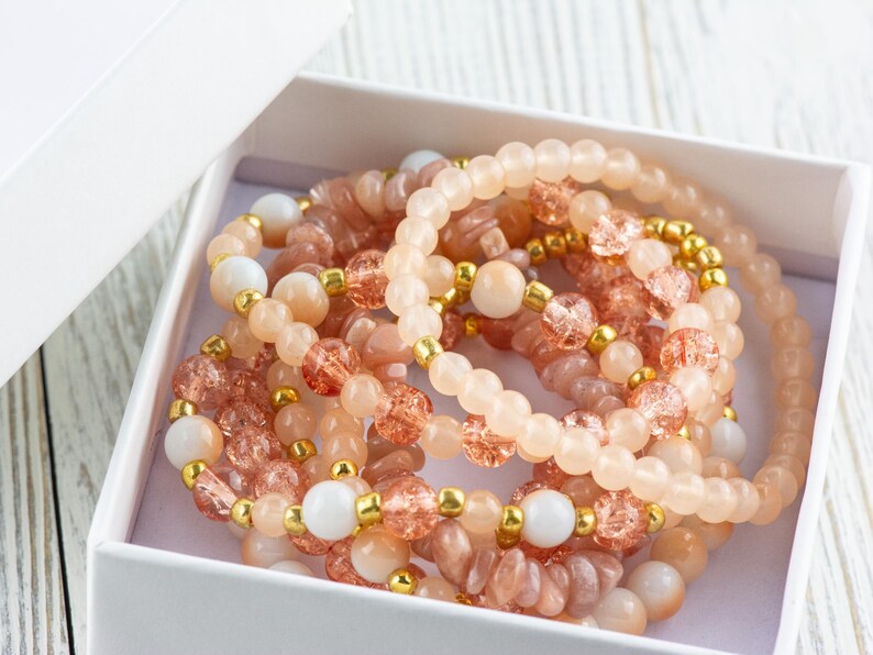 May include: A set of six bracelets made with pink, peach, and white beads. The bracelets are stacked inside a white gift box.