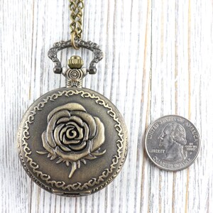 May include: Antique bronze-colored pocket watch with a rose design on the cover. The watch is round and has a decorative border. A quarter is next to the watch for size comparison. The watch hangs from a matching chain.