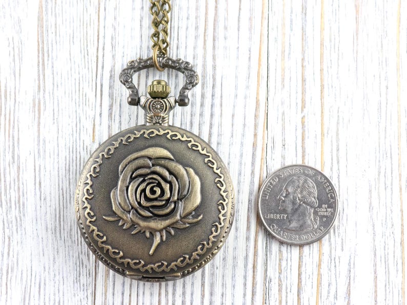 May include: Antique bronze-colored pocket watch with a rose design on the cover. The watch is round and has a decorative border. A quarter is next to the watch for size comparison. The watch hangs from a matching chain.