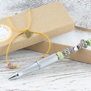K&ouml;nnte beinhalten: Ein silberner Stift mit einem gr&uuml;nen und rosa Perlenmuster. Der Stift befindet sich in einer braunen Schachtel mit einer gelben Schnur und einem Herzanh&auml;nger mit der Aufschrift "Handgefertigt mit Liebe".