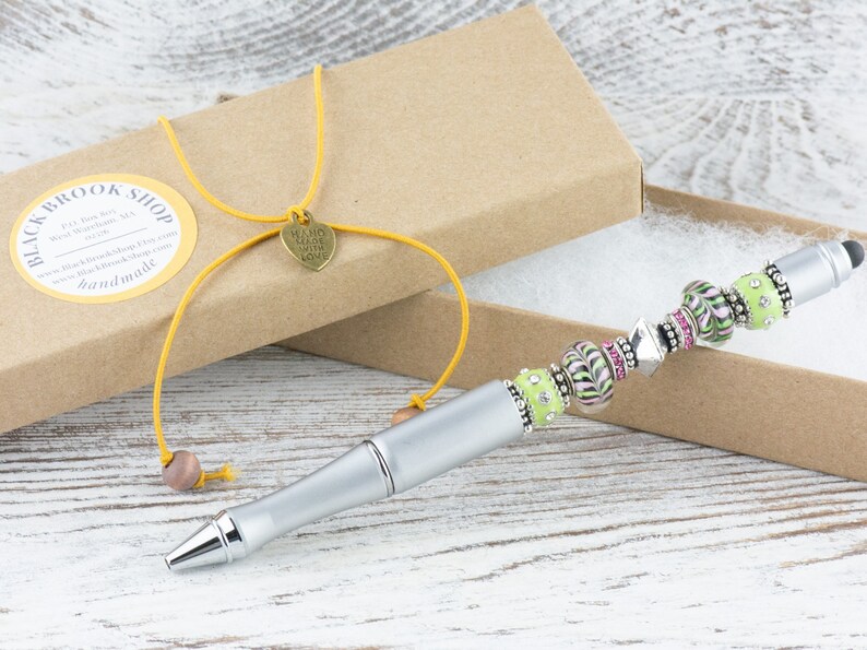 K&ouml;nnte beinhalten: Ein silberner Stift mit einem gr&uuml;nen und rosa Perlenmuster. Der Stift befindet sich in einer braunen Schachtel mit einer gelben Schnur und einem Herzanh&auml;nger mit der Aufschrift "Handgefertigt mit Liebe".