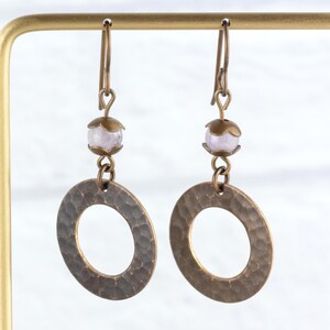 Puede incluir: Un par de pendientes hechos a mano con un dise&ntilde;o de c&iacute;rculo abierto martillado en tono bronce. Cada pendiente tiene una peque&ntilde;a cuenta de color p&uacute;rpura claro y una tapa decorativa en tono bronce. Los pendientes cuelgan de ganchos para las orejas en tono bronce.