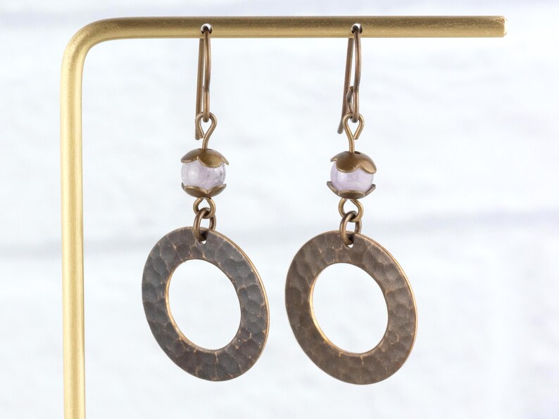 Puede incluir: Un par de pendientes hechos a mano con un dise&ntilde;o de c&iacute;rculo abierto martillado en tono bronce. Cada pendiente tiene una peque&ntilde;a cuenta de color p&uacute;rpura claro y una tapa decorativa en tono bronce. Los pendientes cuelgan de ganchos para las orejas en tono bronce.