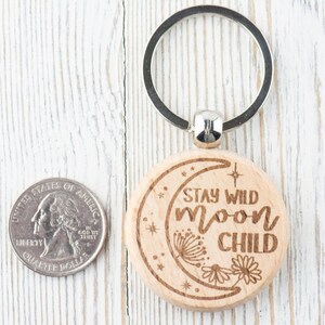 May include: A wooden keychain with a crescent moon design and the text "Stay Wild Moon Child". The keychain is attached to a silver key ring.