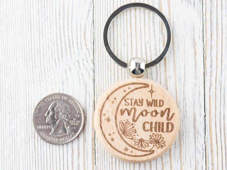 May include: A wooden keychain with a crescent moon design and the text "Stay Wild Moon Child". The keychain is attached to a silver key ring.