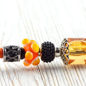May include: A black and orange beaded bracelet with various decorative beads. The beads include a silver bead with black accents, an orange and yellow bead, a black textured bead, and a large amber-colored bead with a silver filigree setting.