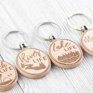 K&ouml;nnte beinhalten: Holz-Schl&uuml;sselanh&auml;nger mit silbernen Schl&uuml;sselringen. Jeder Schl&uuml;sselanh&auml;nger ist rund und mit einem Naturmotiv und den Worten "Beach Life", "River Life", "Lake Life" und "Mountain Life" graviert. Die Schl&uuml;sselanh&auml;nger sind aus hellem Holz.