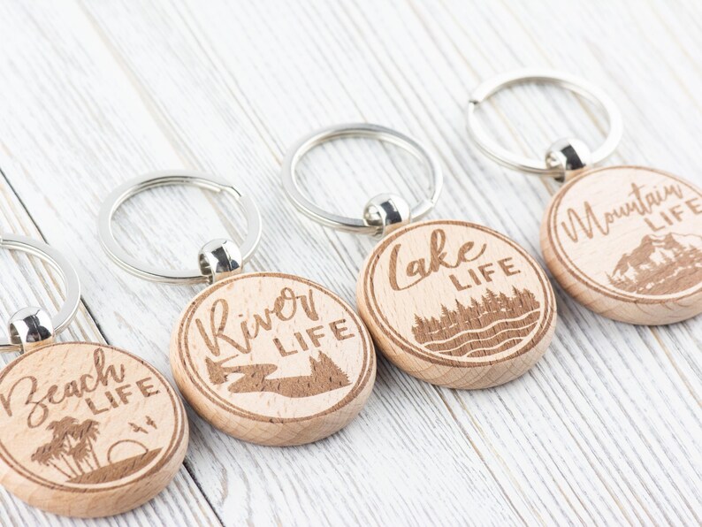 K&ouml;nnte beinhalten: Holz-Schl&uuml;sselanh&auml;nger mit silbernen Schl&uuml;sselringen. Jeder Schl&uuml;sselanh&auml;nger ist rund und mit einem Naturmotiv und den Worten "Beach Life", "River Life", "Lake Life" und "Mountain Life" graviert. Die Schl&uuml;sselanh&auml;nger sind aus hellem Holz.