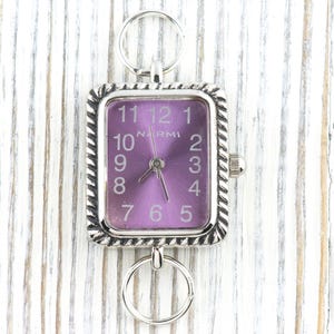 以下が含まれることがあります： 紫色の文字盤とロープのような装飾的な縁取りが付いた、銀色の長方形の時計。時計には、上部と下部にアタッチメント用のリングが付いています。文字盤にはブランド名「NARMI」と1から12までの数字が表示されています。
