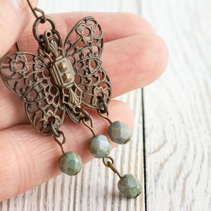 May include: Antique bronze-toned butterfly earrings with intricate filigree wings. The butterfly design features a central rectangular element and is adorned with three dangling, faceted, pale green beads. The earrings are held in a hand against a white wood background.