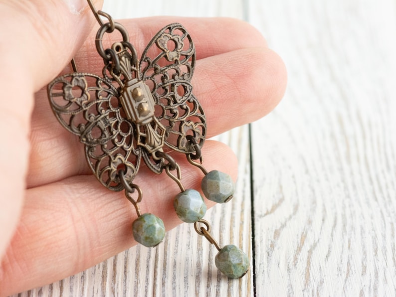 May include: Antique bronze-toned butterfly earrings with intricate filigree wings. The butterfly design features a central rectangular element and is adorned with three dangling, faceted, pale green beads. The earrings are held in a hand against a white wood background.