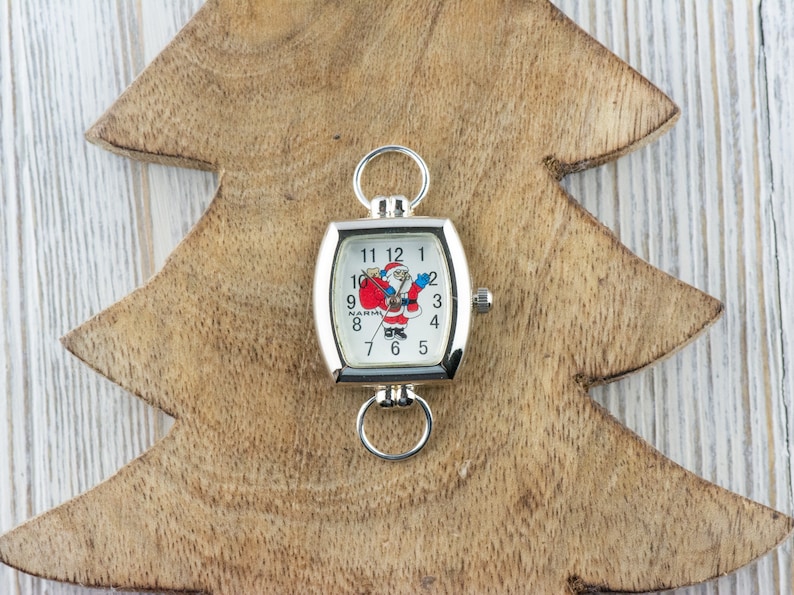 May include: A silver square watch with a white face and a Santa Claus design. The watch is on a wooden Christmas tree cutout.