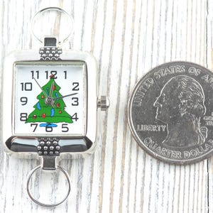 Puede incluir: Un reloj de bolsillo plateado con un &aacute;rbol de Navidad en la esfera blanca. El reloj presenta un dise&ntilde;o de &aacute;rbol de Navidad verde con adornos rojos y azules. Una moneda de 25 centavos estadounidense est&aacute; al lado del reloj.
