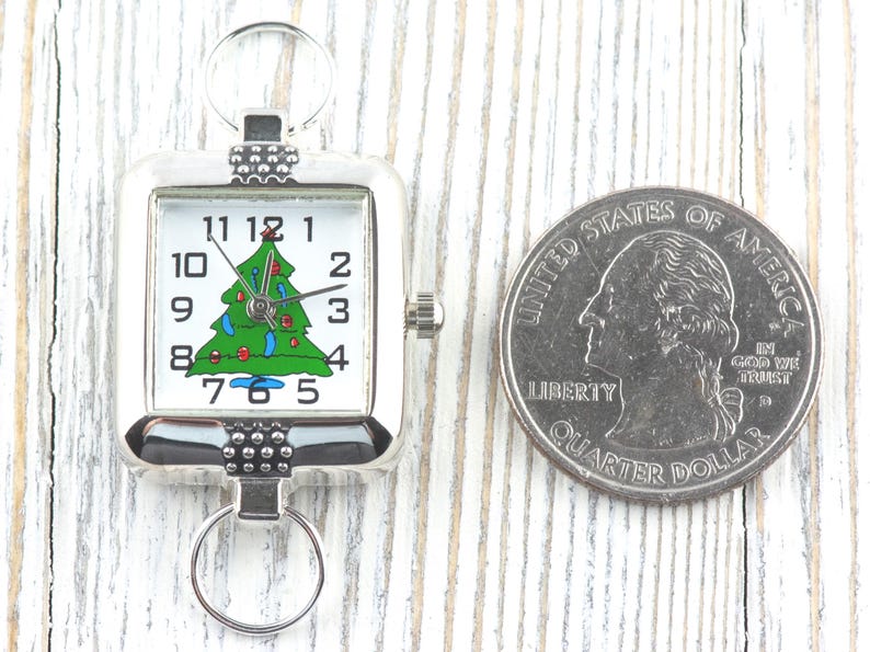 Puede incluir: Un reloj de bolsillo plateado con un &aacute;rbol de Navidad en la esfera blanca. El reloj presenta un dise&ntilde;o de &aacute;rbol de Navidad verde con adornos rojos y azules. Una moneda de 25 centavos estadounidense est&aacute; al lado del reloj.