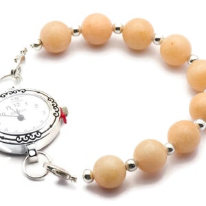 Puede incluir: Un reloj plateado con una esfera blanca y n&uacute;meros negros est&aacute; unido a una pulsera hecha de cuentas de color naranja claro.