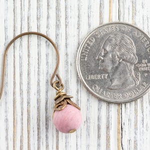 K&ouml;nnte beinhalten: Ein einzelner Ohrring mit einer rosa Perle und bronzefarbenen Metallakzenten. Der Ohrring befindet sich neben einer US-Vierteldollarm&uuml;nze zum Gr&ouml;&szlig;envergleich. Der Ohrring hat einen Hakenverschluss.