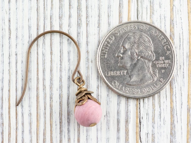 K&ouml;nnte beinhalten: Ein einzelner Ohrring mit einer rosa Perle und bronzefarbenen Metallakzenten. Der Ohrring befindet sich neben einer US-Vierteldollarm&uuml;nze zum Gr&ouml;&szlig;envergleich. Der Ohrring hat einen Hakenverschluss.