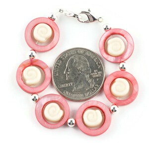 Pu&ograve; includere: Un braccialetto di conchiglie rosa e bianche con una chiusura argentata. Il braccialetto &egrave; circa delle dimensioni di una moneta da 25 centesimi di dollaro americano.