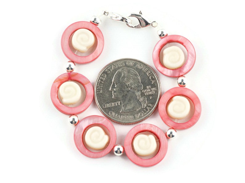 Pu&ograve; includere: Un braccialetto di conchiglie rosa e bianche con una chiusura argentata. Il braccialetto &egrave; circa delle dimensioni di una moneta da 25 centesimi di dollaro americano.