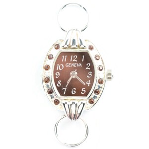K&ouml;nnte beinhalten: Eine silberne Uhr mit braunem Zifferblatt und silbernem Armband. Die Uhr hat ein rundes Zifferblatt mit r&ouml;mischen Ziffern f&uuml;r die Stunden und einen kleinen Sekundenzeiger. Das Armband besteht aus zwei silbernen Ringen, die durch eine silberne Stange verbunden sind. Die Uhr ist mit kleinen braunen Strasssteinen um das Zifferblatt herum verziert.