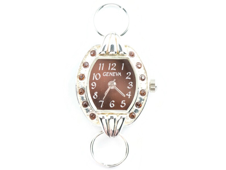 K&ouml;nnte beinhalten: Eine silberne Uhr mit braunem Zifferblatt und silbernem Armband. Die Uhr hat ein rundes Zifferblatt mit r&ouml;mischen Ziffern f&uuml;r die Stunden und einen kleinen Sekundenzeiger. Das Armband besteht aus zwei silbernen Ringen, die durch eine silberne Stange verbunden sind. Die Uhr ist mit kleinen braunen Strasssteinen um das Zifferblatt herum verziert.