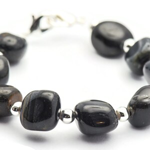 K&ouml;nnte beinhalten: Ein Armband aus schwarzen Edelsteinen mit silbernen Perlen und einem Verschluss. Das Armband besteht aus glatten, abgerundeten Steinen.