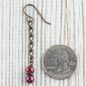 Puede incluir: Un solo pendiente colgante con una cadena y un gancho de color cobre oscuro. Dos cuentas de color p&uacute;rpura oscuro est&aacute;n unidas a la parte inferior de la cadena. El pendiente est&aacute; al lado de una moneda de 25 centavos estadounidense para comparar el tama&ntilde;o.