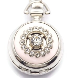 May include: A silver pocket watch with a pink face and a crystal embellishment in the center. The watch has a decorative chain attached to the top.