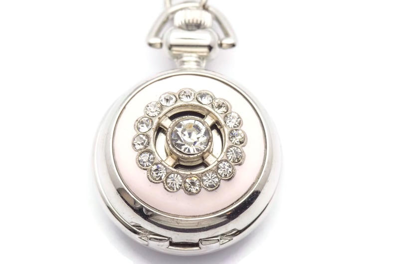 May include: A silver pocket watch with a pink face and a crystal embellishment in the center. The watch has a decorative chain attached to the top.