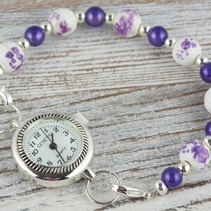 May include: A silver pocket watch with a white face and black numbers is attached to a purple and white beaded bracelet. The watch face reads "GENEVA QUARTZ".