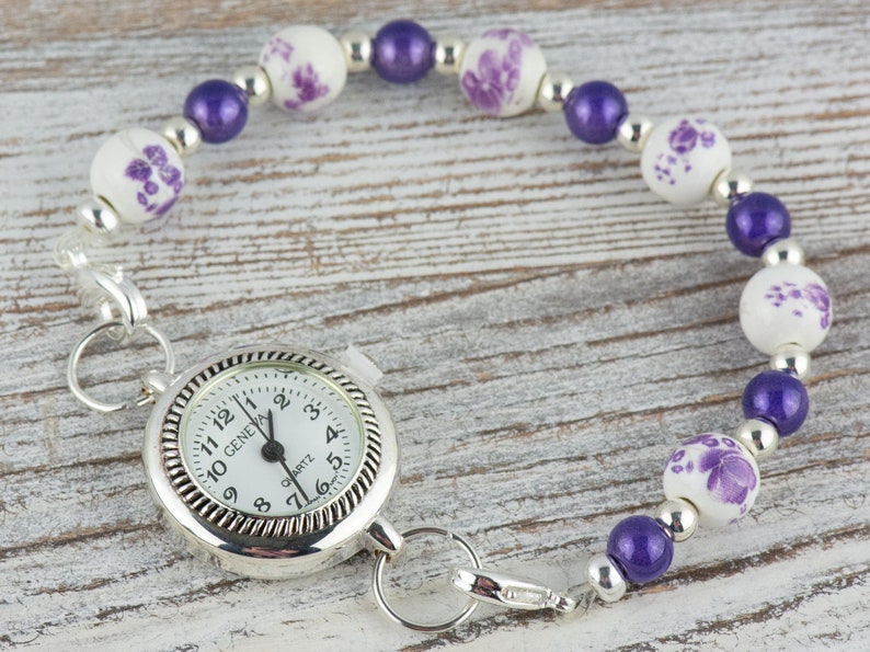 May include: A silver pocket watch with a white face and black numbers is attached to a purple and white beaded bracelet. The watch face reads "GENEVA QUARTZ".