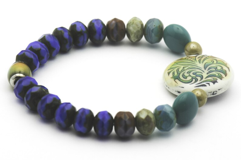 Puede incluir: Un brazalete de cuentas azules, verdes y marrones con un dije de cer&aacute;mica blanca con un dise&ntilde;o floral verde.