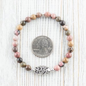 Puede incluir: Un brazalete de piedras preciosas rosa, marr&oacute;n y verde con un dije de plata. El brazalete es aproximadamente del tama&ntilde;o de una moneda de 25 centavos.