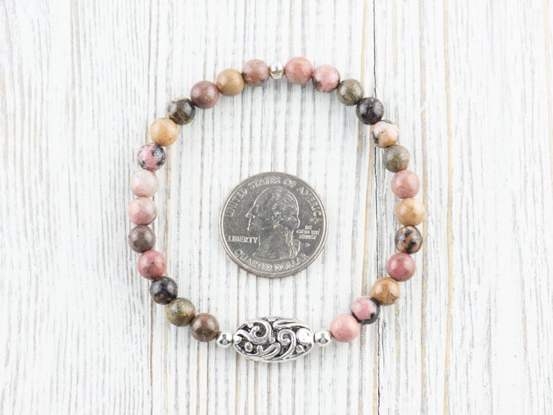 Puede incluir: Un brazalete de piedras preciosas rosa, marr&oacute;n y verde con un dije de plata. El brazalete es aproximadamente del tama&ntilde;o de una moneda de 25 centavos.