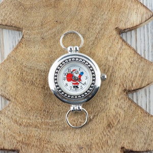 Puede incluir: Un reloj de bolsillo plateado con una esfera blanca y un dise&ntilde;o de Pap&aacute; Noel. El reloj tiene un borde con cuentas y una cadena plateada.