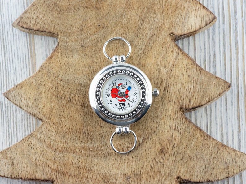 Puede incluir: Un reloj de bolsillo plateado con una esfera blanca y un dise&ntilde;o de Pap&aacute; Noel. El reloj tiene un borde con cuentas y una cadena plateada.