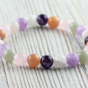 May include: A beaded bracelet featuring a variety of colorful gemstones. The bracelet includes round beads in shades of purple, pink, orange, white, and green. The bracelet is displayed on a white wooden surface.
