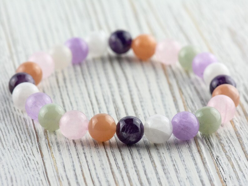 May include: A beaded bracelet featuring a variety of colorful gemstones. The bracelet includes round beads in shades of purple, pink, orange, white, and green. The bracelet is displayed on a white wooden surface.